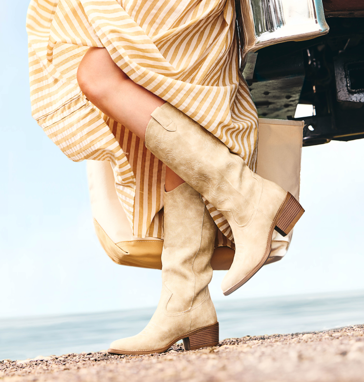 Woman wearing a yellow and white striped dress and beige suede knee-high boots with stacked wooden heels, sitting on the edge of a vehicle near the beach.